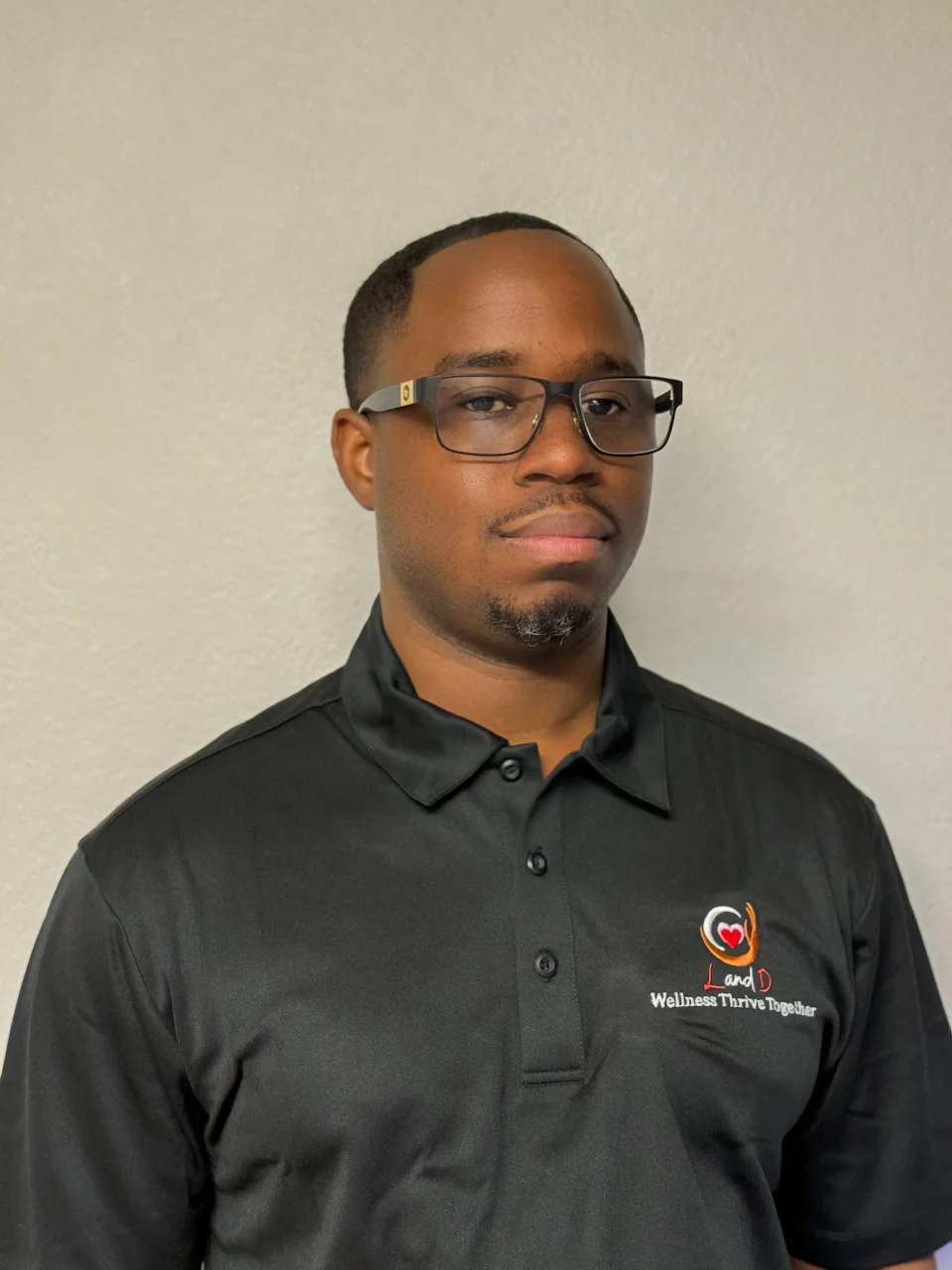 A man with short hair, wearing glasses and a black polo shirt with a logo on the chest standing against a plain wall.