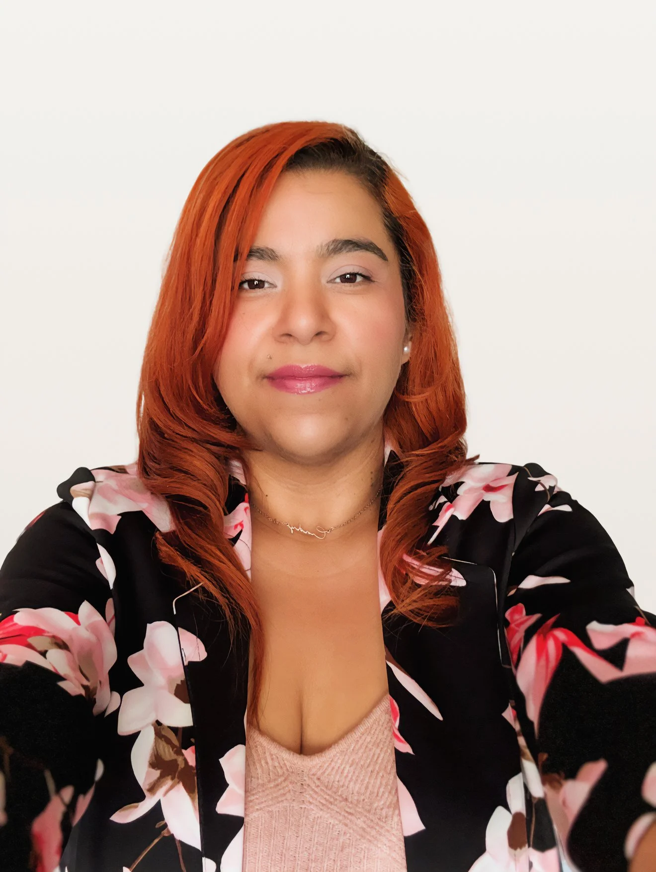 A woman with long, wavy auburn hair and light skin taking a selfie against a plain white background, wearing a black floral blazer with pink and beige flowers, a pink top, and a delicate necklace.