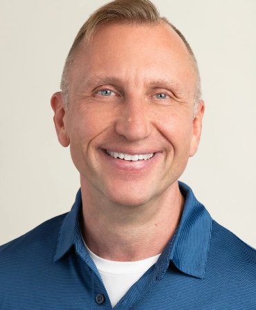 Portrait of a smiling middle-aged man with short blond hair wearing a blue button-up shirt and a white undershirt against a plain light background.