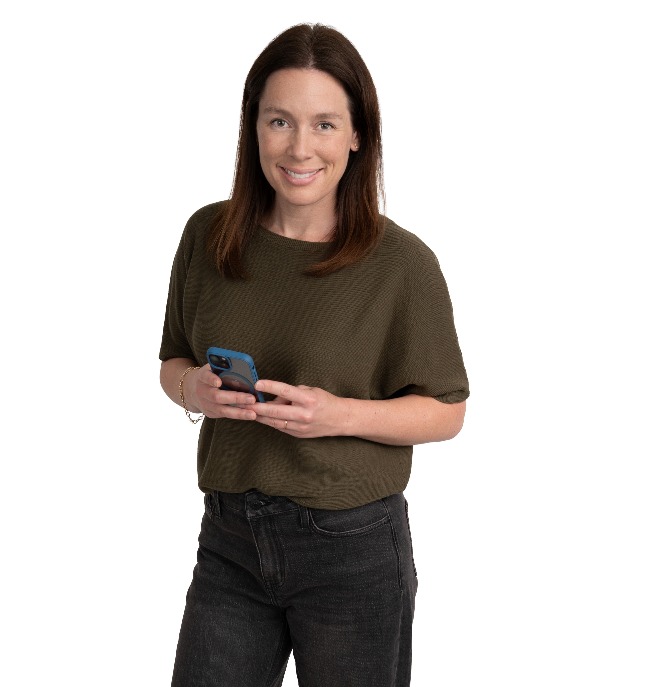 A woman with shoulder-length brown hair wearing a dark green top and black jeans, holding a smartphone, smiling at the camera.