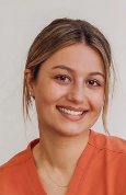 A young woman with light brown hair and a bright smile wearing an orange top, standing against a plain background.