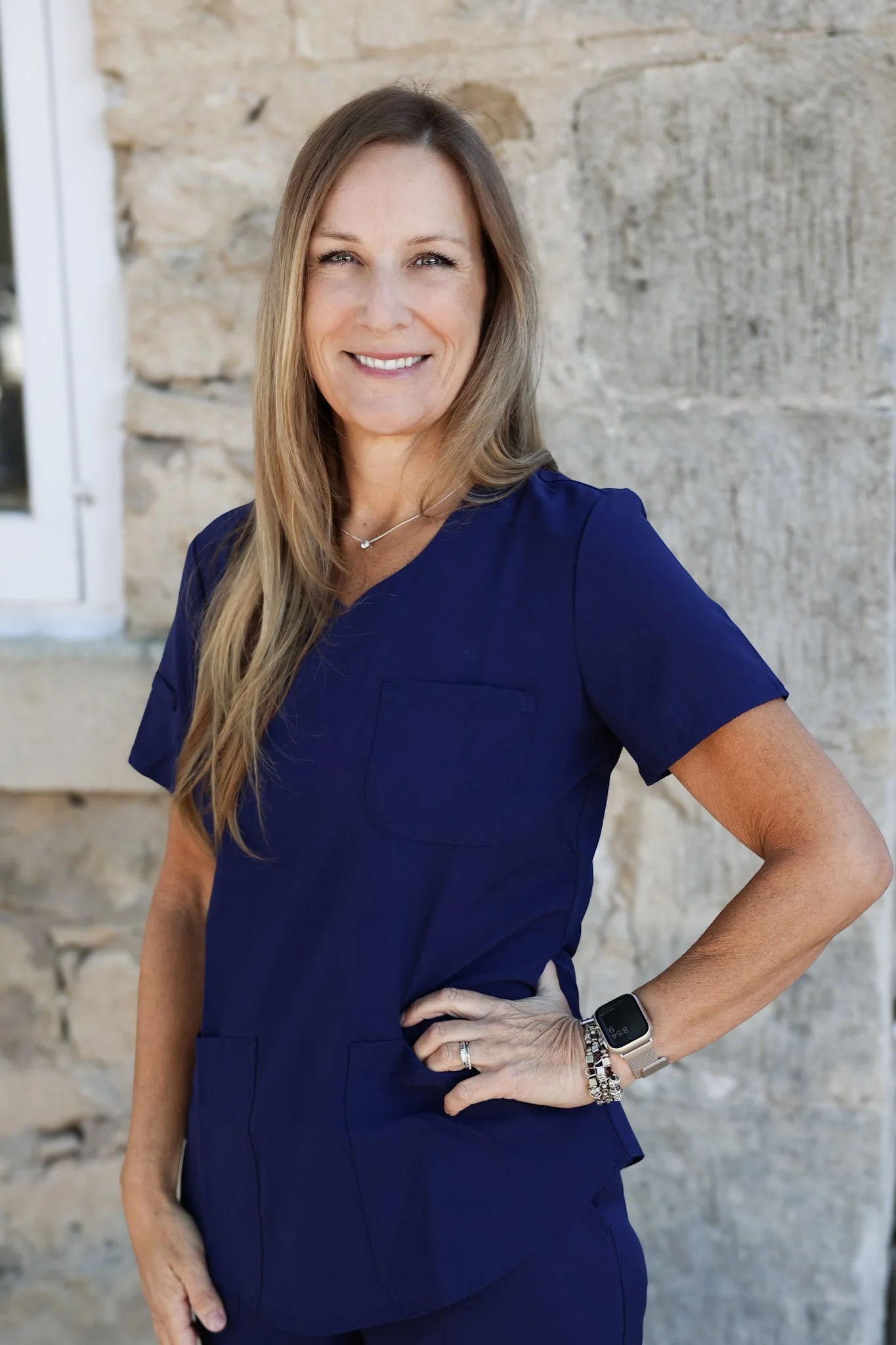 A woman with long, light brown hair smiling, wearing navy scrubs, a smartwatch, and jewelry, standing outdoors against a stone wall.