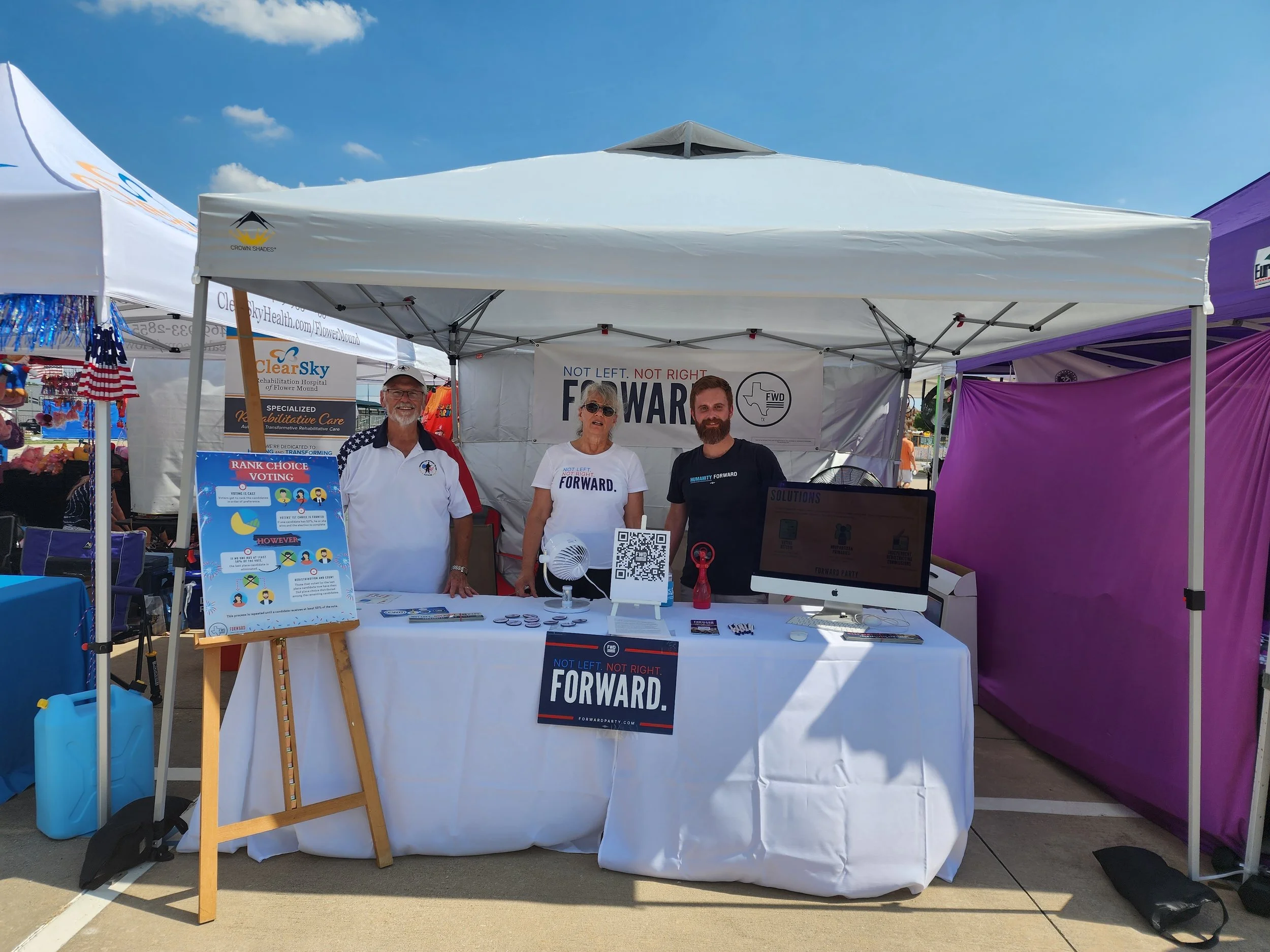 Three people standing behind a booth with a banner that reads 'NOT LEFT. NOT RIGHT. FORWARD.' at an outdoor event under a white canopy. The booth has a computer, QR code, and informational materials on the table.