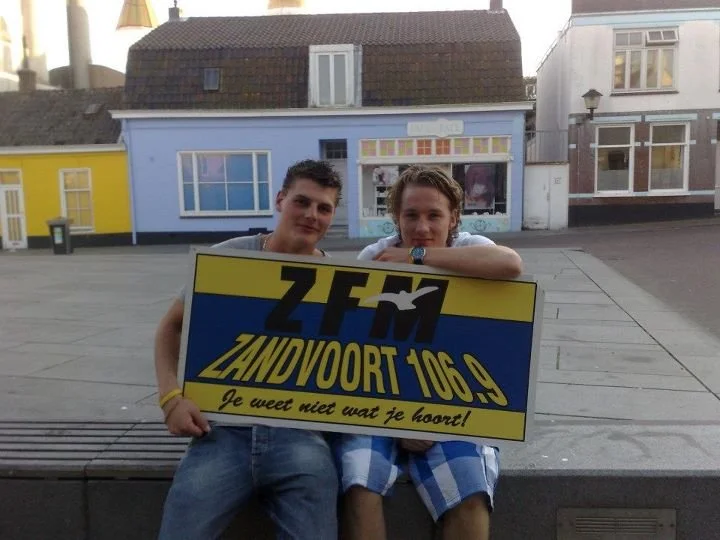 Twee jongens zitten op een bankje in een stad, met een bord van een lokale radiozender ZFM Zandvoort 106.9, en lopen naast kleurrijke gebouwen.