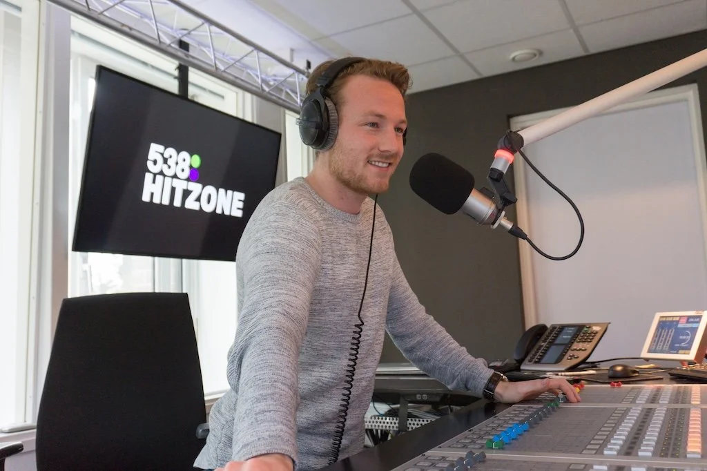 Man in radiostudio met koptelefoon voor microfoon, met radio- en audiovoorzieningen, zittend en werkend.