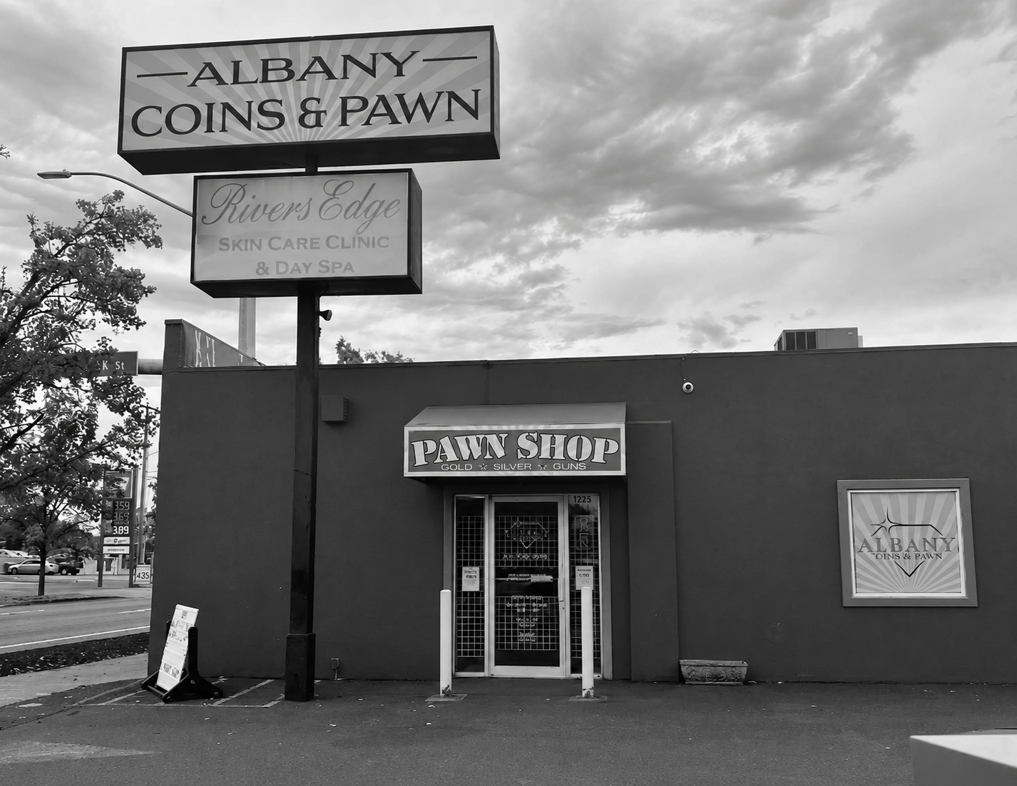 Black and white photo of a building with a sign that reads 'Pawn Shop', a smaller sign above the entrance reads 'Rivers Edge Skin Care Clinic & Day Spa', and a large sign on a pole reads 'Albany Coins & Pawn'. There are trees and cars visible in the background.
