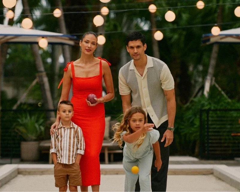 A family of four playing bocce ball outdoors with lush green surroundings. The woman is wearing a bright red dress and the man is wearing a casual short-sleeved button-up shirt. There is a young boy and girl and the girl is tossing a yellow ball.