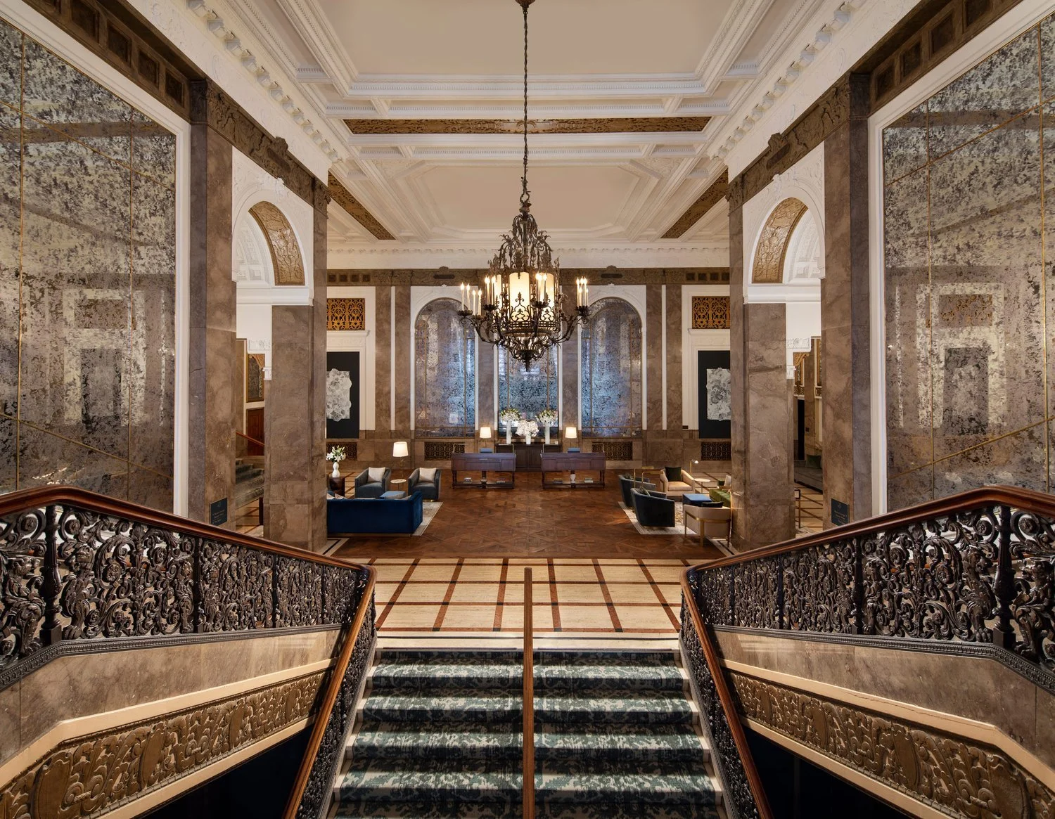 The grand hotel lobby of the Beacon Grand with a sweeping staircase, ornate iron railings, and marble flooring. A large chandelier hangs from the ceiling, and there are comfortable-looking seating areas with plush couches and armchairs.