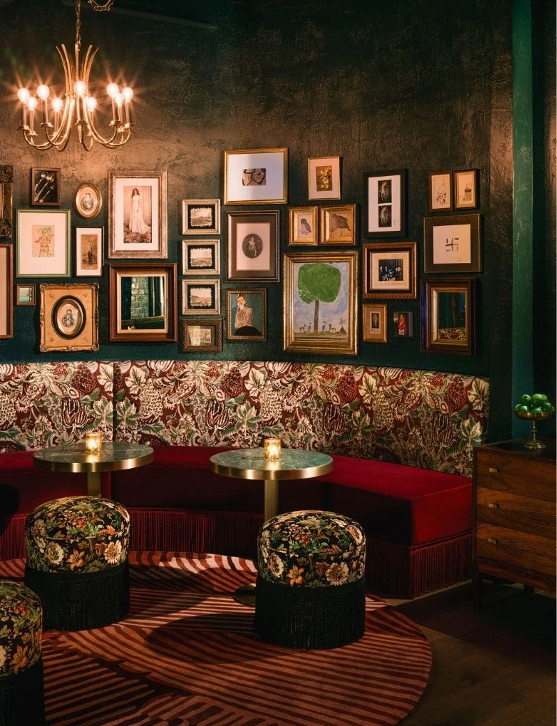 A cozy lounge area at Starlite, The Beacon Grand. The walls are deep green and adorned with an eclectic collection of framed artwork. The lounge features plush red velvet seating, patterned ottomans, and a brass table lamp.