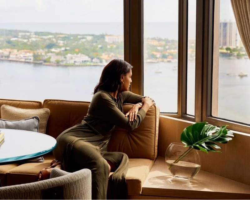 A woman in a dark brown dress sits on a brown leather sofa looking out a high-rise window. The scenery outdoors shows the ocean and a bit of land with buildings in the distance.