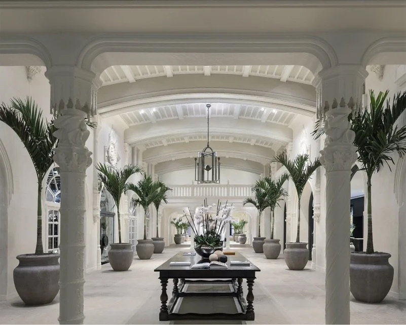 A grand, white marble lobby with tall columns and arched doorways at The Boca Raton. Lush greenery in large planters lines the hallway, with a central table displaying books and flowers. Sunlight streams in through the arched windows.