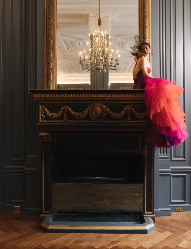 A woman in a bright pink and orange ball gown sits on the mantel of a large, ornate fireplace. The mantel is black with gold details. A large gold-framed mirror sits above it and shows the reflection of a chandelier. The walls are a deep blue.
