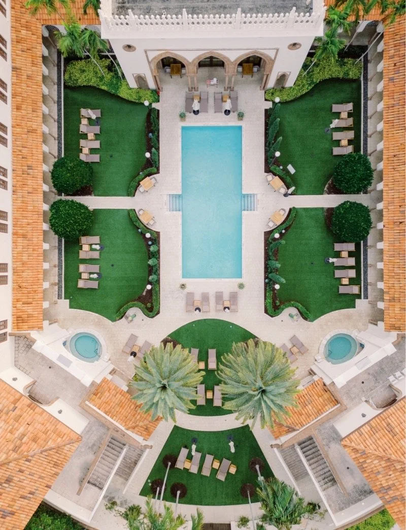 An aerial view of a courtyard at The Boca Raton resort. The courtyard features a rectangular pool surrounded by lush green lawns, palm trees, and sun loungers. The building architecture is Mediterranean-inspired with red tiled roofs.