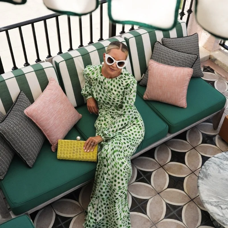 A woman wearing large white sunglasses and a green dotted dress sits on green and white lounge furniture.