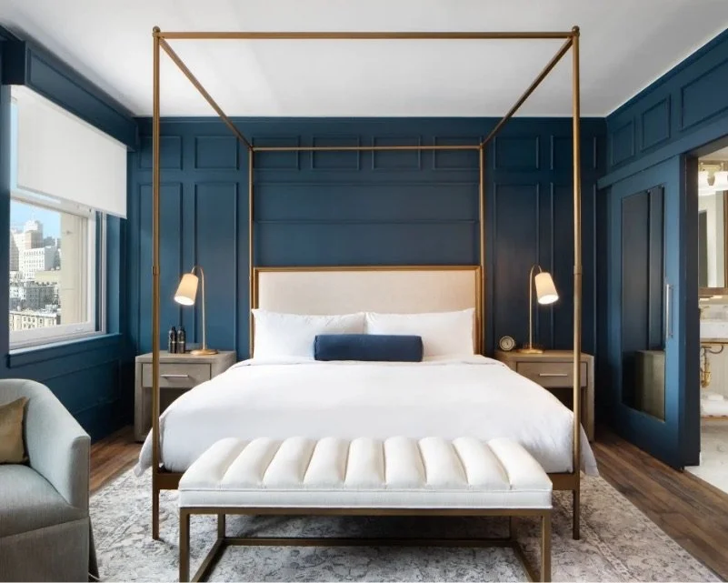 A luxurious guest room at The Beacon Grand, featuring a four-poster gold bed frame with crisp white linens. The room has deep blue walls with gold accents, a plush white bench, and armchair. Soft natural light fills the room.
