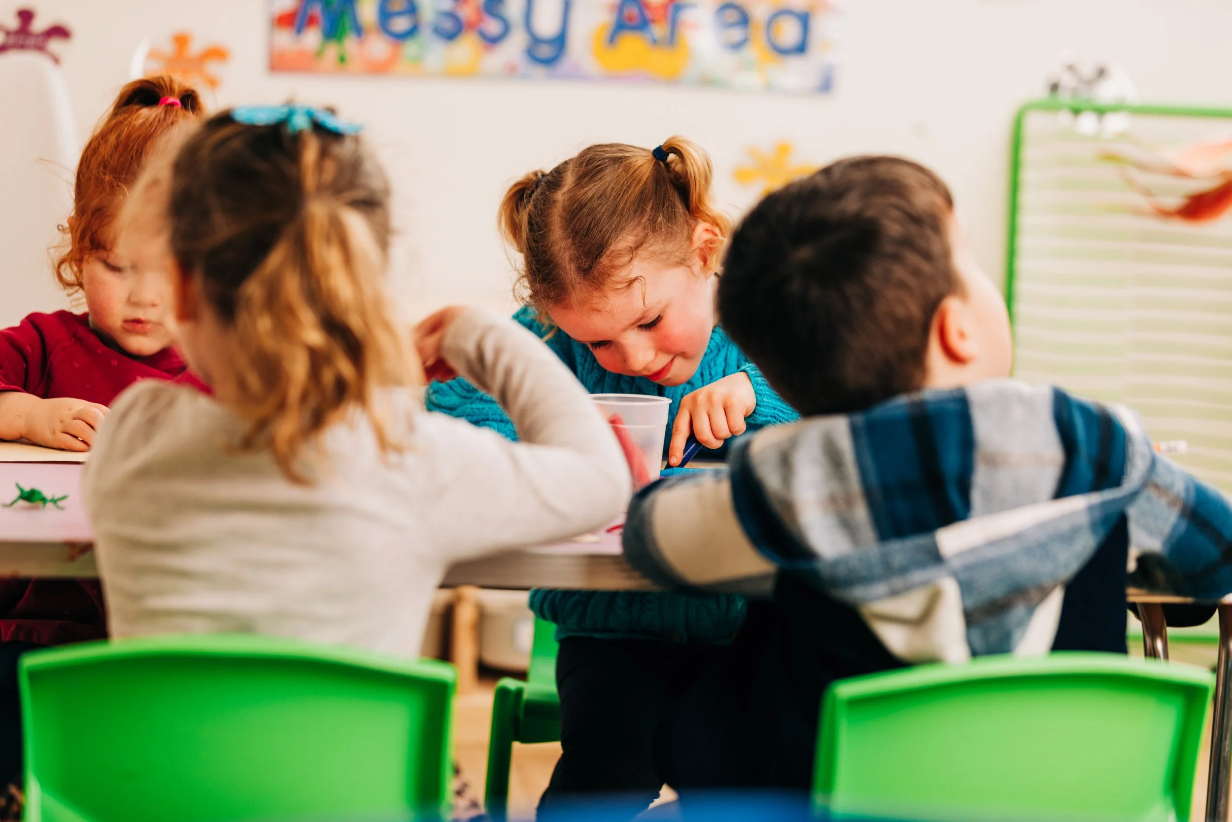 Young children sitting at desks in Make Waves Nursery, Bideford, engaging in an activity, with colourful chairs and learning posters on the wall.