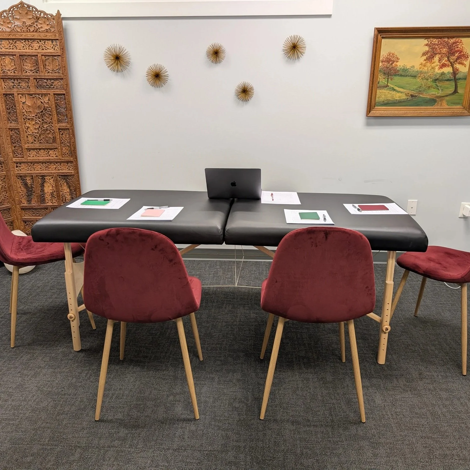 Image of a massage table with 4 chairs around the table. There is a computer, papers, small notebooks, and pens for 3 participants to sit, along with Laura as the facilitator.