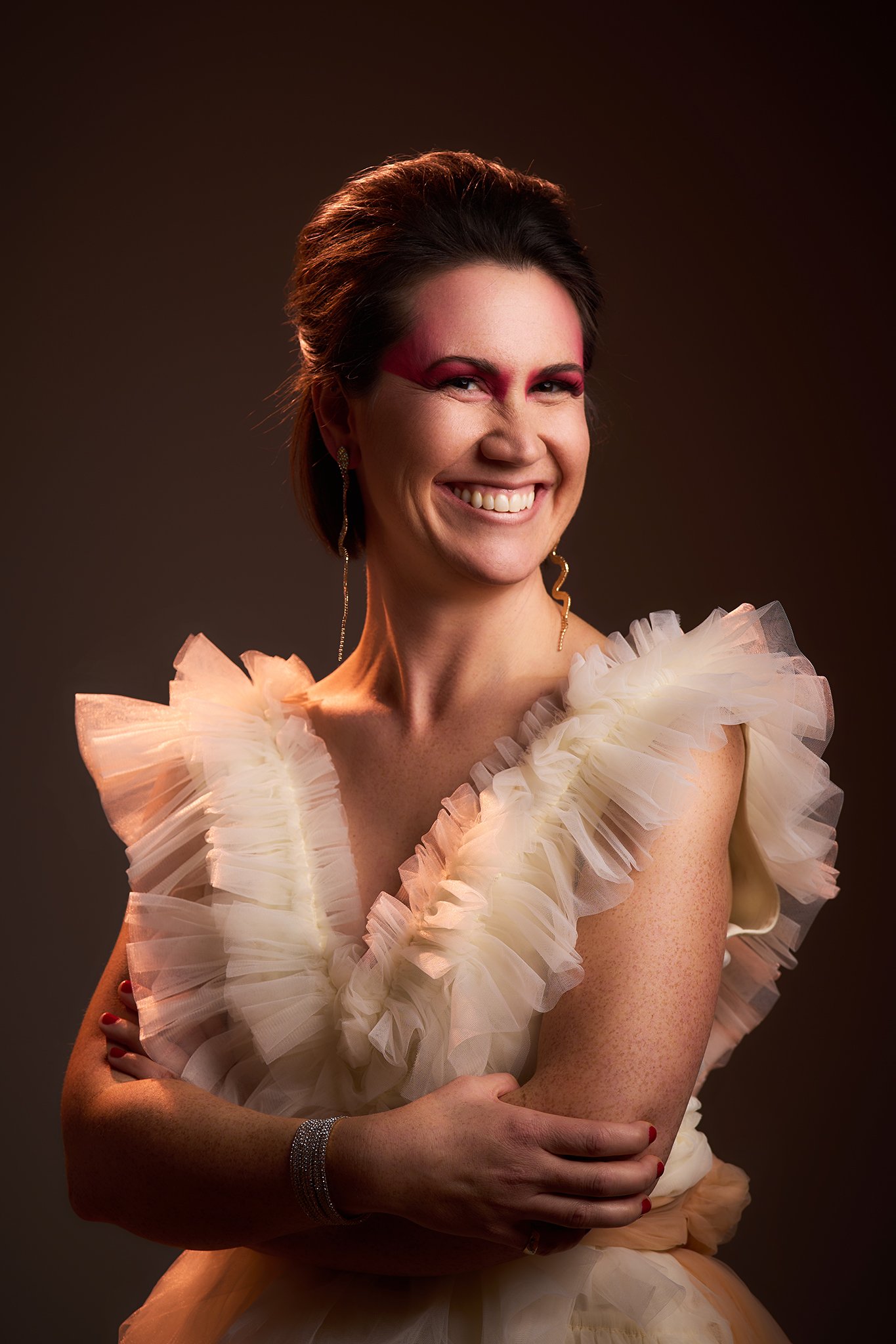 A woman smiling, with short dark hair, wearing a cream-colored dress with ruffled details and gold earrings, standing against a dark background.