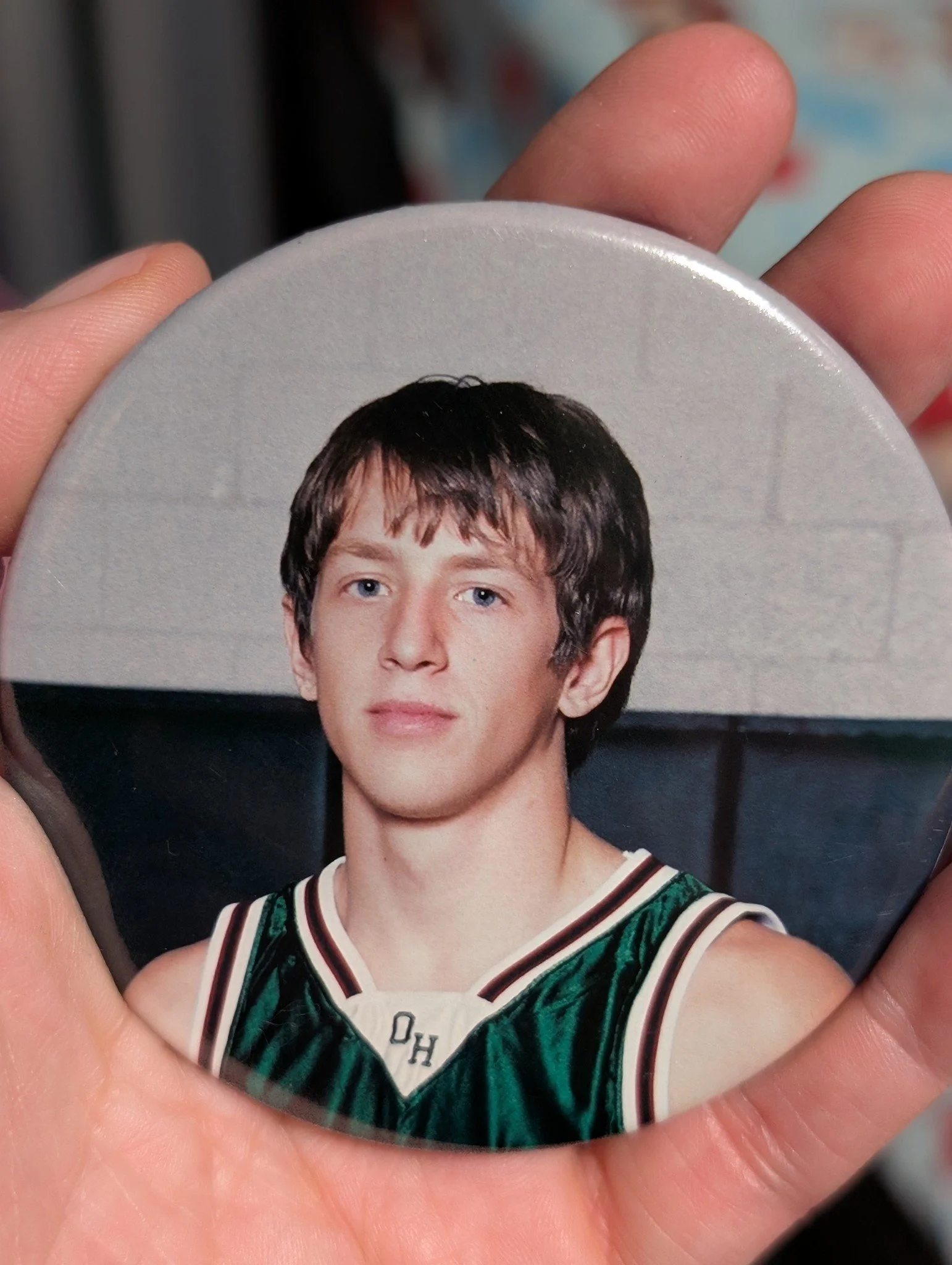 Close-up of a person holding a round button featuring a young man with dark, slightly wavy hair, blue eyes, and wearing a green basketball jersey with white and red trim.