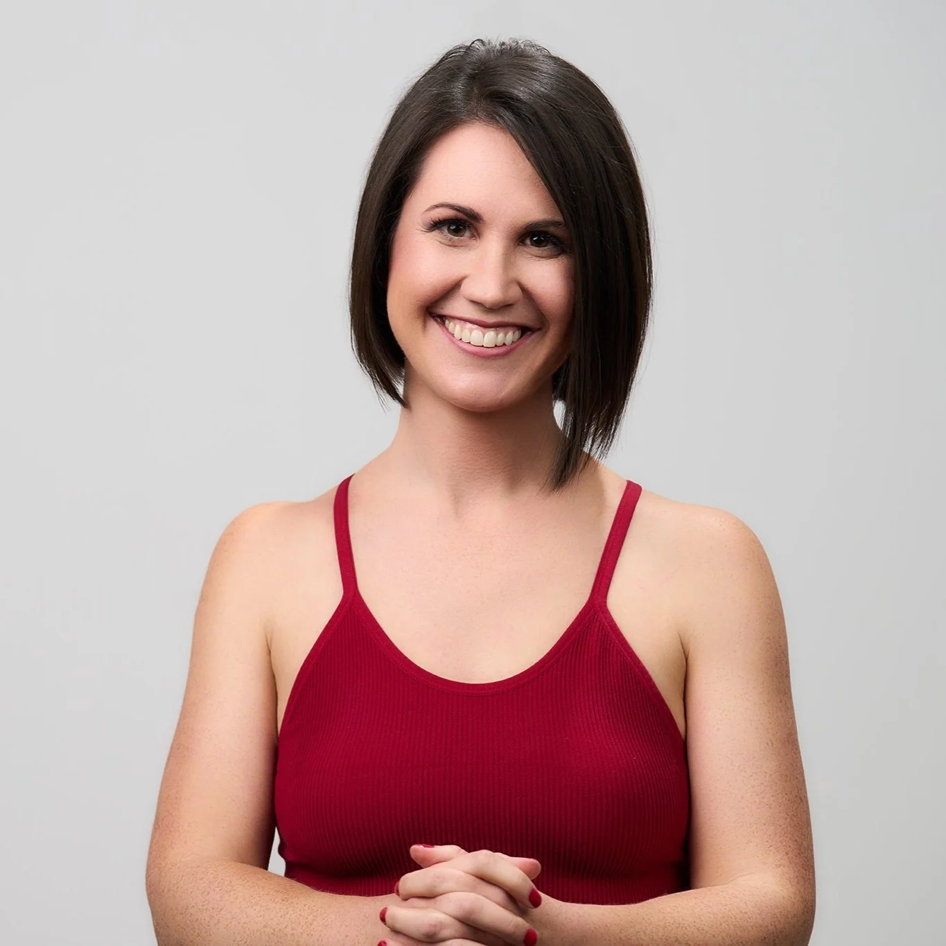 A woman with shoulder-length dark brown hair, wearing a red tank top, smiling and standing against a plain light gray background.