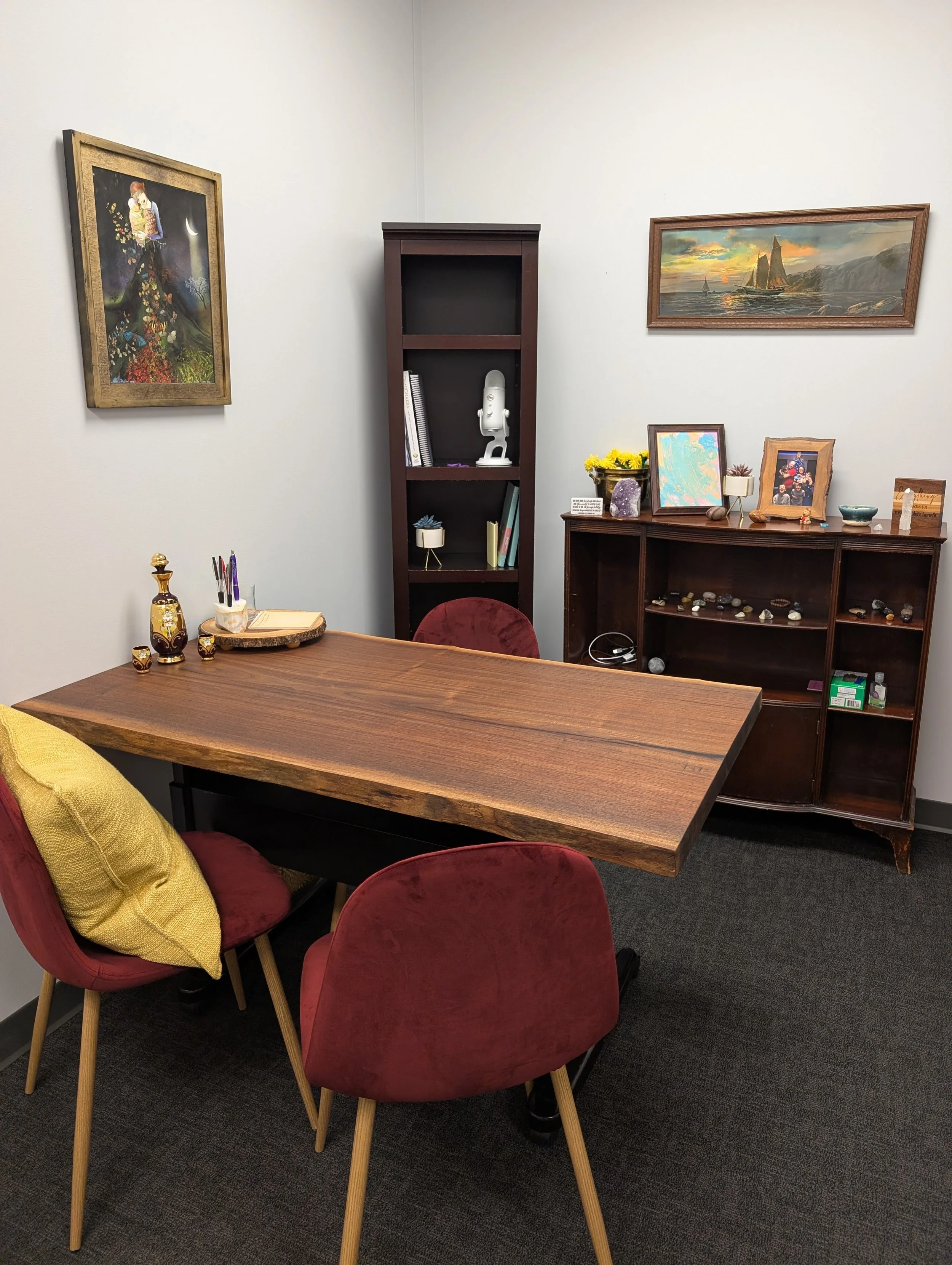 Image of the far side of the office including 2 large wall images, a bookshelf, wooden desk table with 3 chairs and a chest with pictures and crystals
