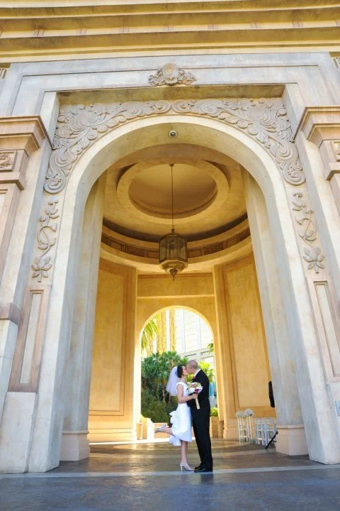 A bride and groom standing close together under a large ornate marble archway, with the bride holding a bouquet, in a sunny outdoor setting.