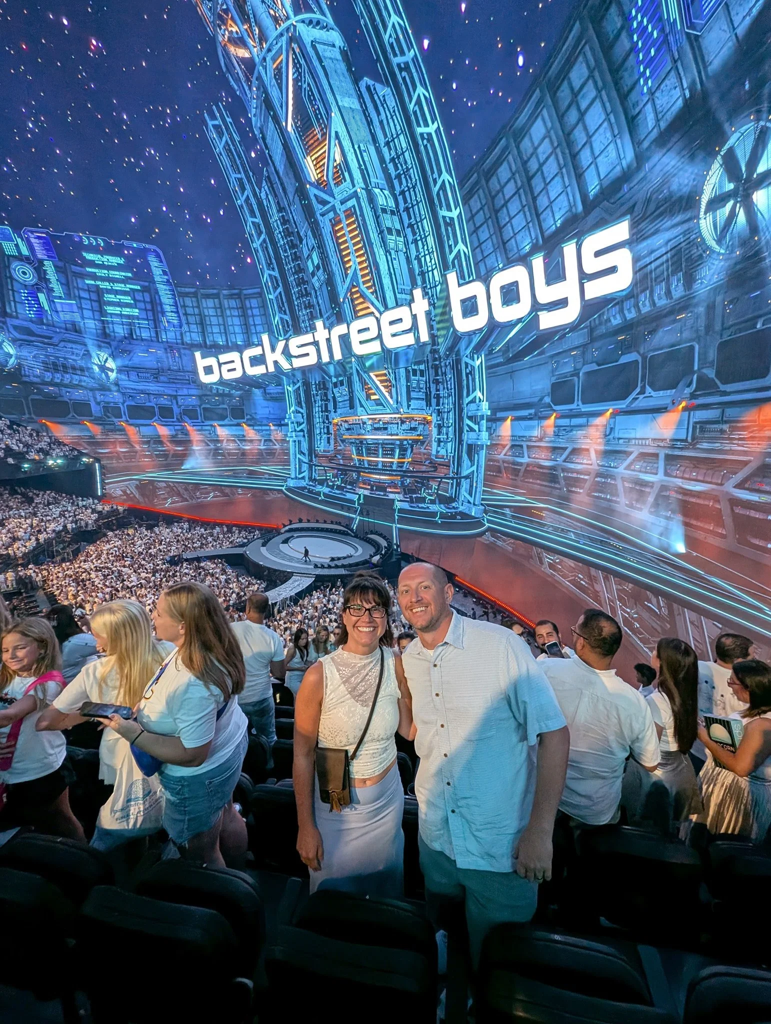 People attending an event inside a futuristic, brightly lit venue with a large sign that reads 'backstreet boys'. The venue features high-tech decor with neon lights and digital displays, and there is a large crowd of spectators.
