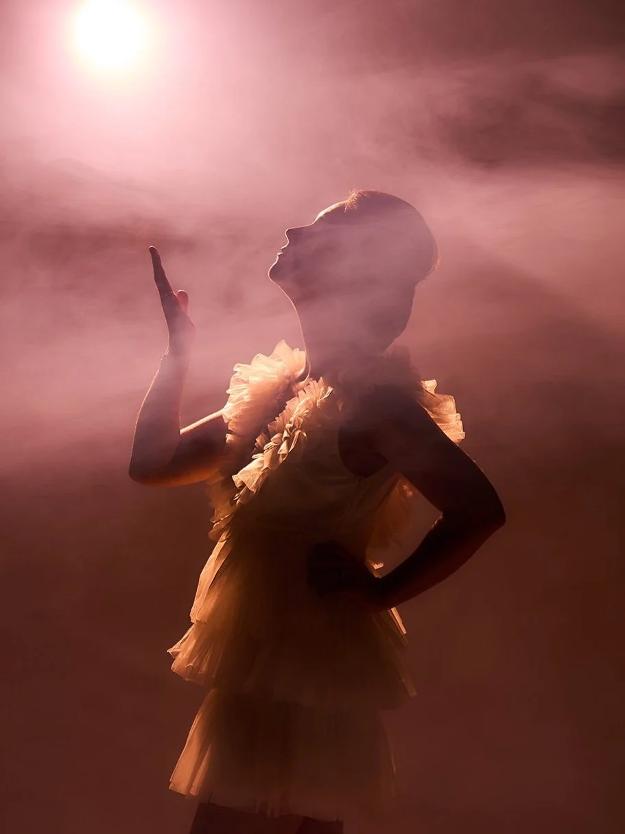 Silhouette of a woman in a tutu dress with puffed sleeves, raising her right hand with her index finger pointing up against a backlit, smoky background.