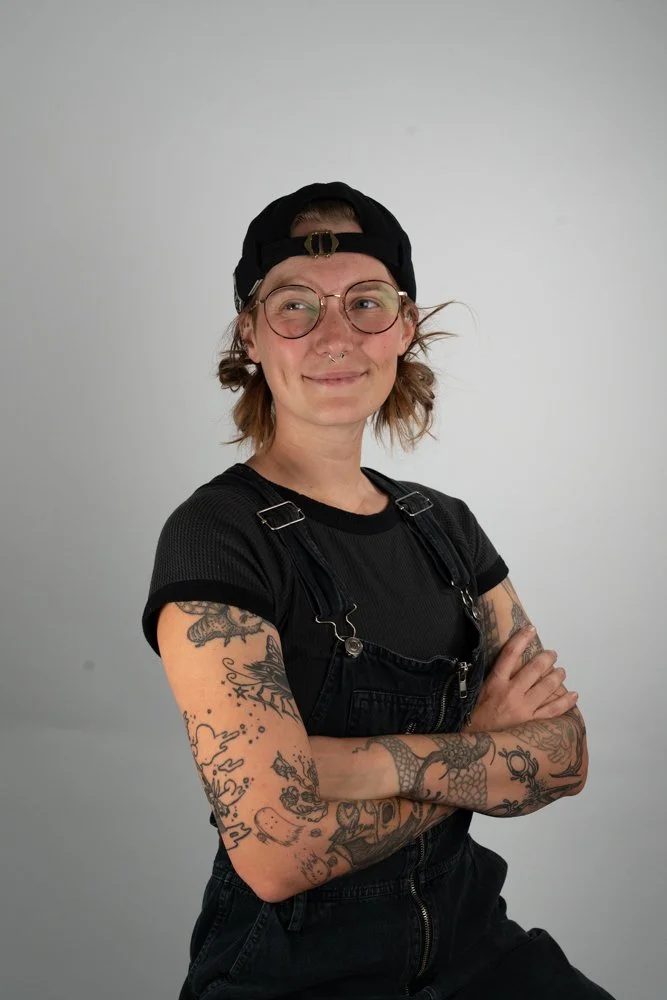 Person with glasses and tattoos on arms, wearing black overalls, a black t-shirt, and a black baseball cap, standing with arms crossed against a plain white background.