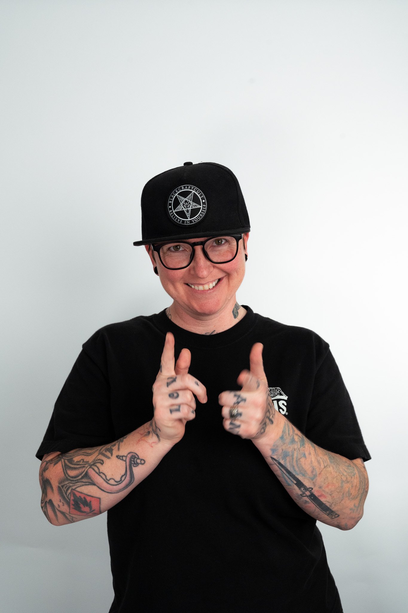 Person smiling and pointing with both hands, wearing glasses, a black hat with a logo, and a black t-shirt with a logo, against a plain light background.