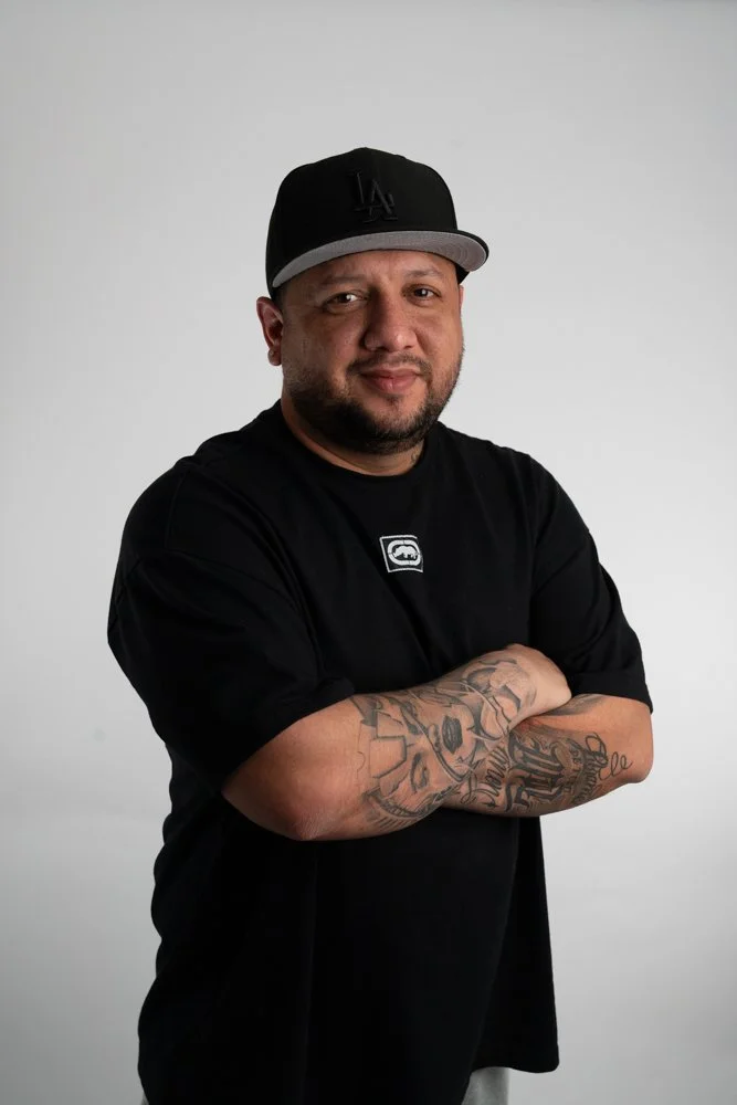 Man with closely shaved beard wearing a black cap and black shirt with tattoos on his arms, standing with arms crossed against a plain light gray background.