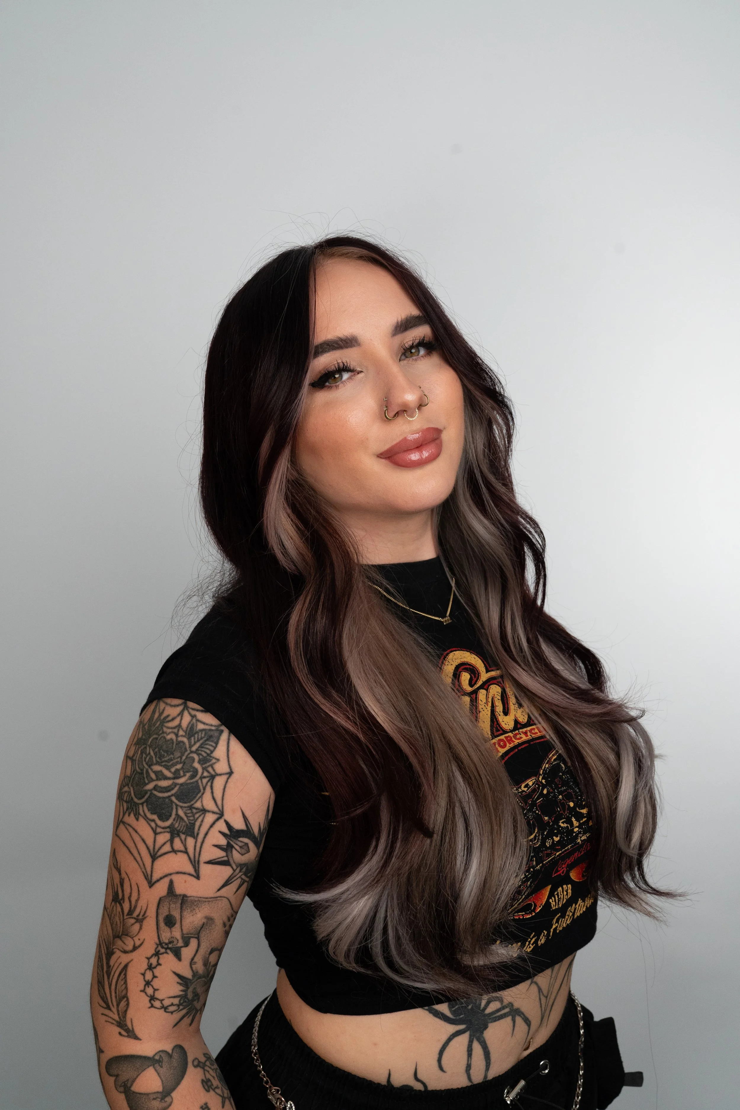 A young woman with long wavy hair featuring dark and gray tones, tattoos on her arm, and wearing a black t-shirt with a graphic design stands against a plain white background.