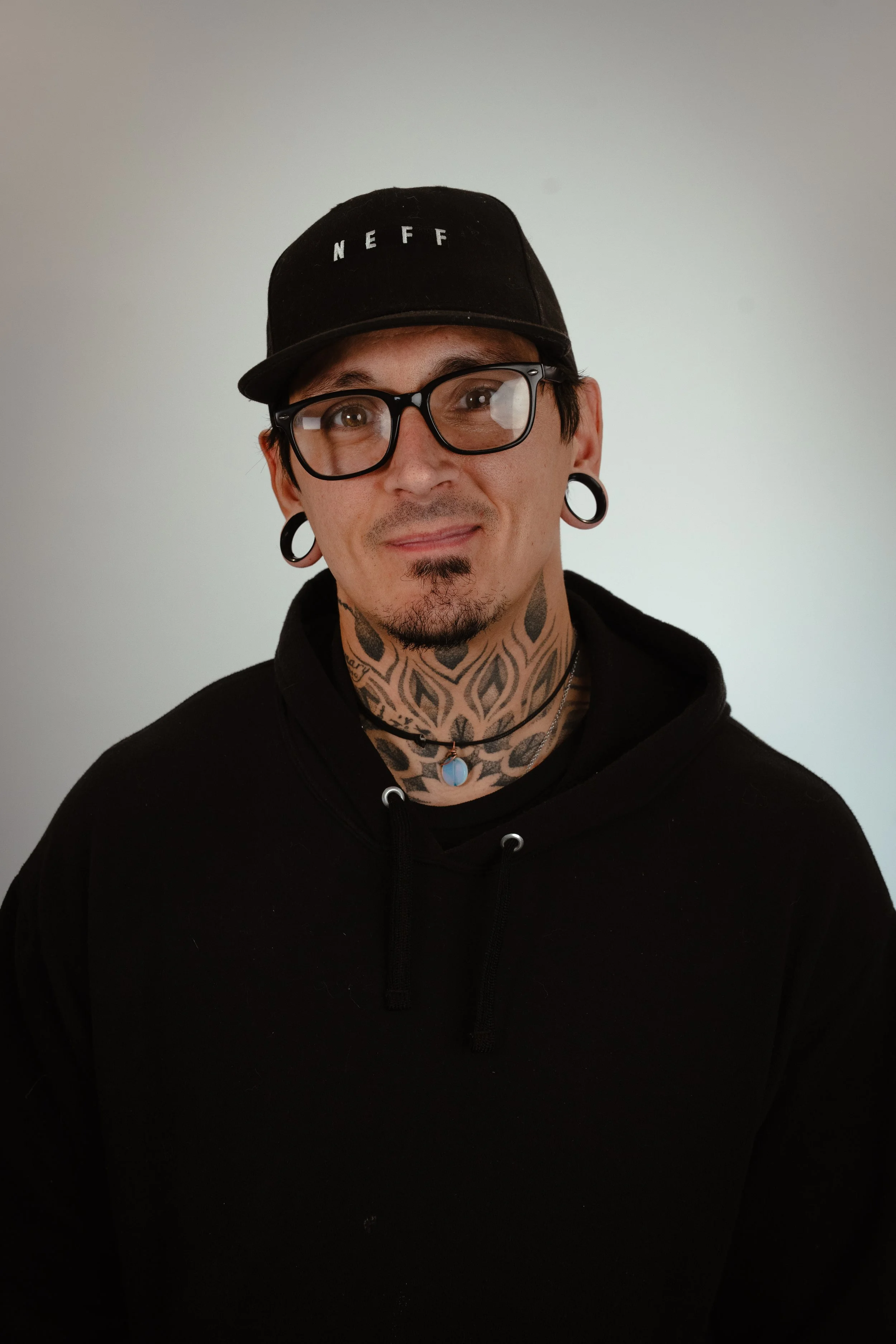 A man with glasses, tattoos on his neck, wearing a black hoodie, cap, and large earrings, standing against a plain wall.