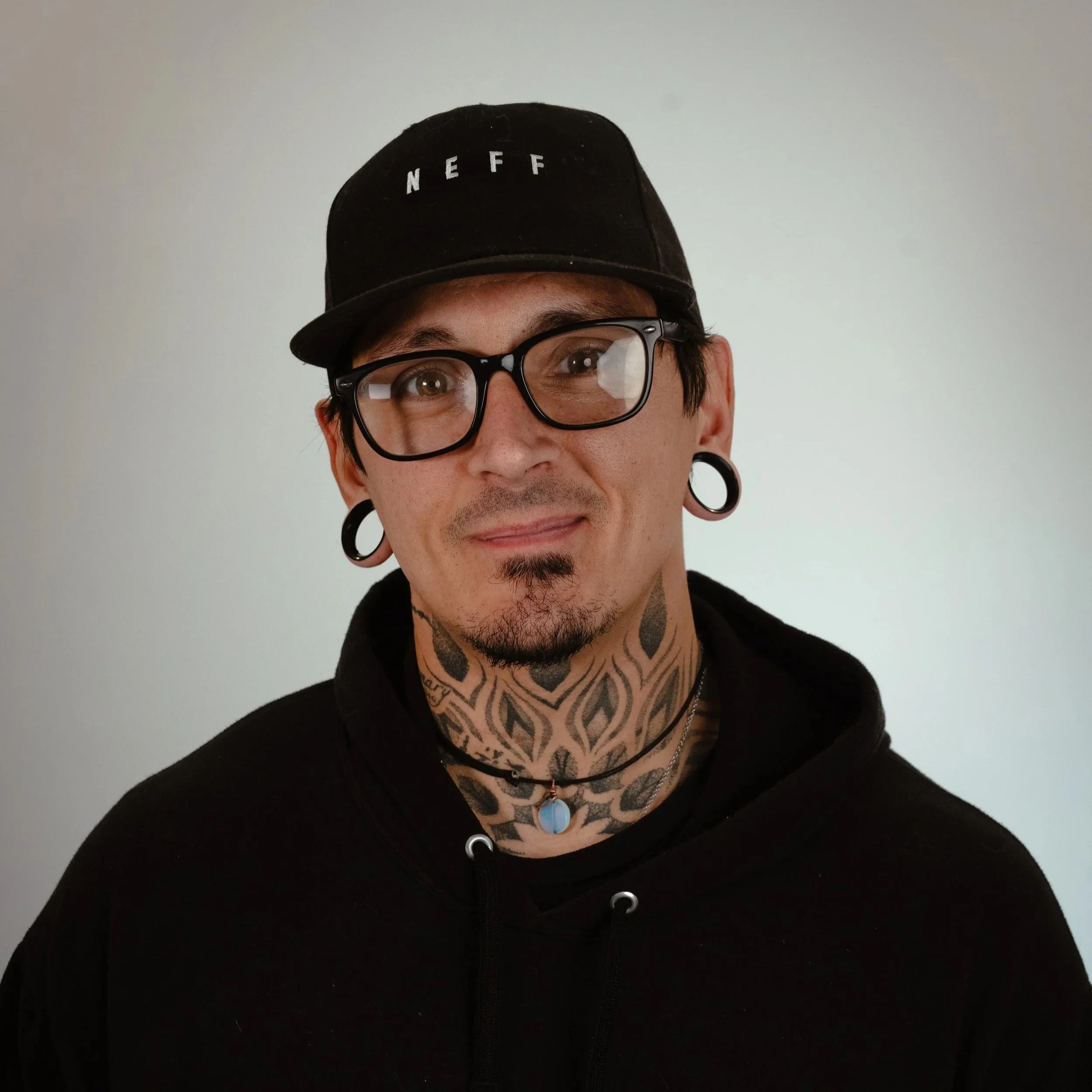 A man with tattoos on his neck, wearing glasses, earrings, and a black cap and hoodie, smiling at the camera.