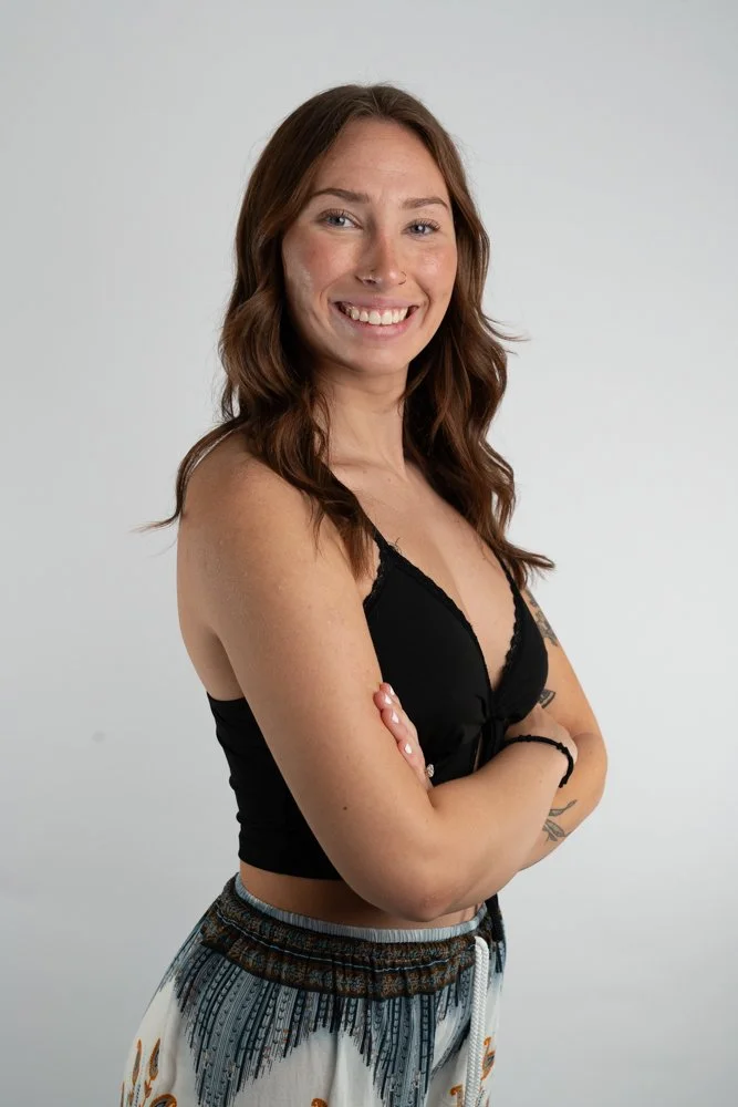 Portrait of a smiling woman with brown hair, arms crossed, wearing a black crop top and patterned shorts, standing against a plain light grey background.