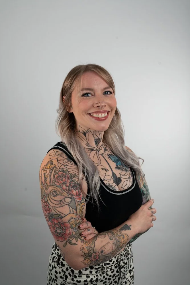 Woman with blonde hair and tattoos on her arms and chest, wearing a black tank top and leopard print pants, smiling and standing against a plain light gray background.