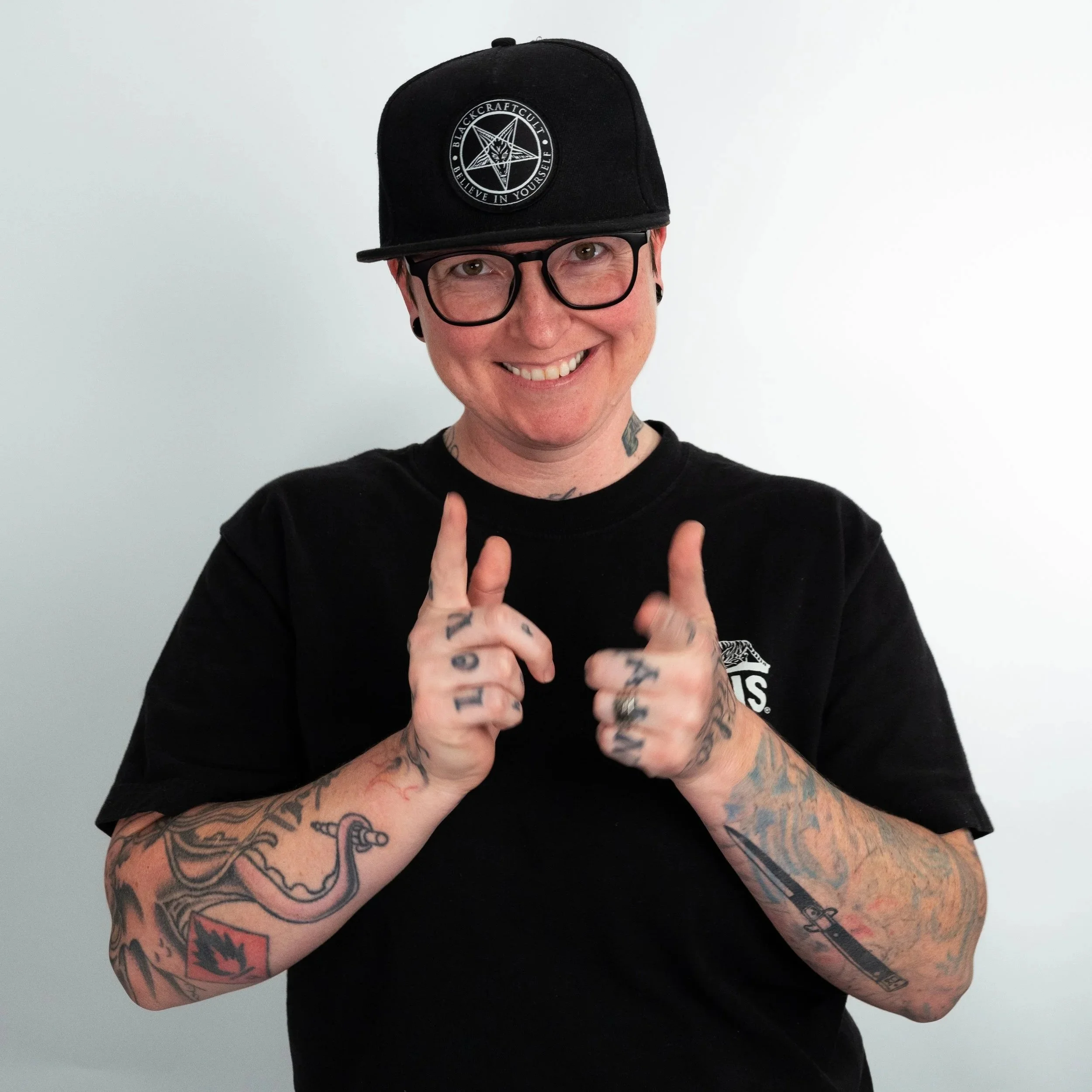 A person with short brown hair, wearing glasses, a black T-shirt, and a red and black plaid shirt, standing against a plain white background.