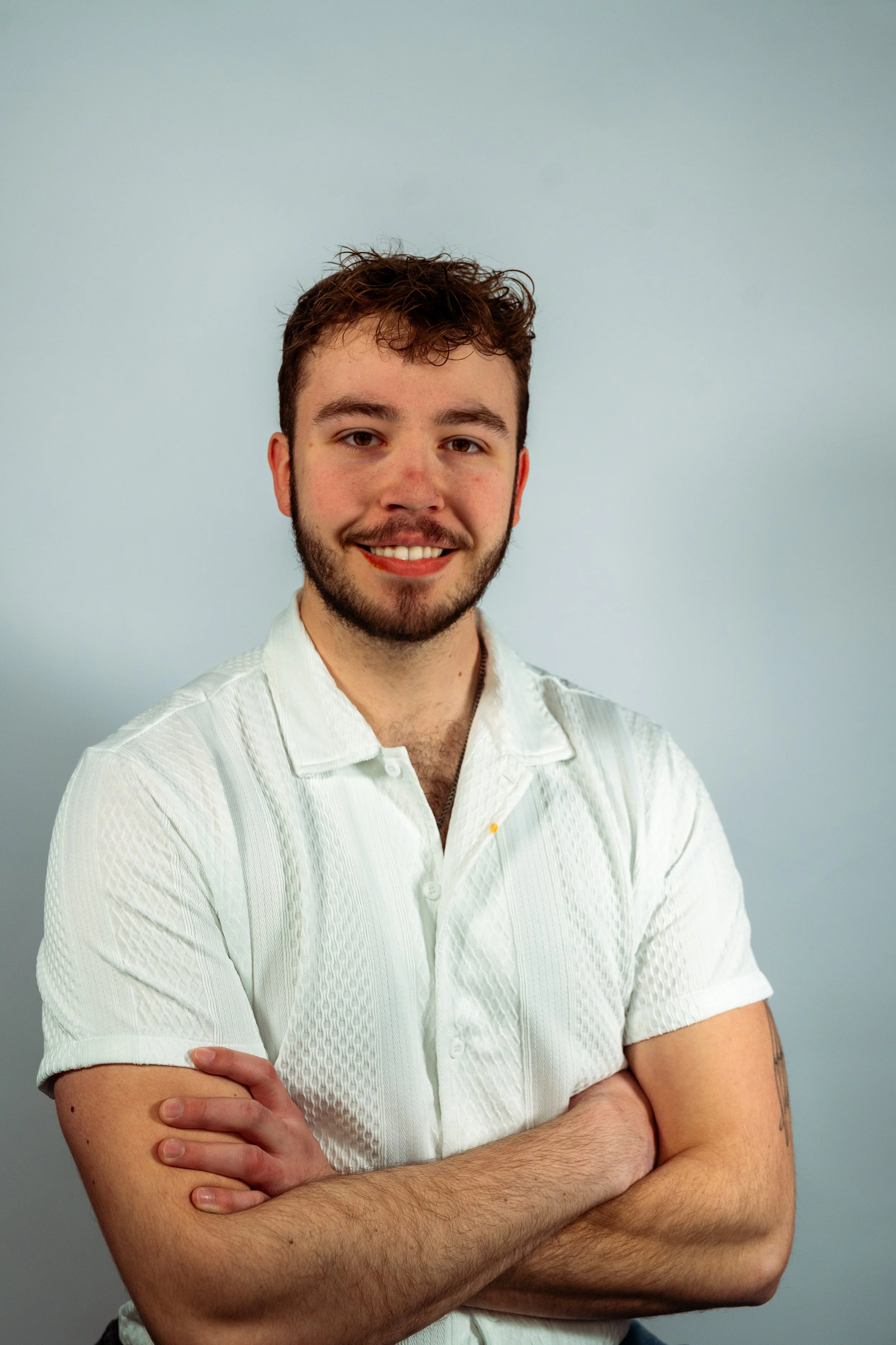 A man with facial hair and a white shirt, crossing his arms, smiling