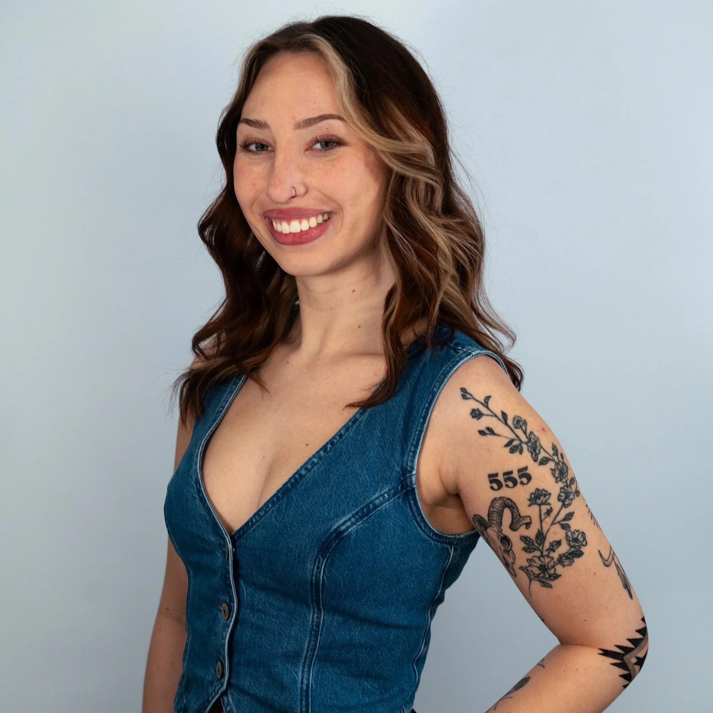A young woman with long, wavy brown hair and light skin smiling, wearing a black spaghetti strap top, standing against a plain light gray background.