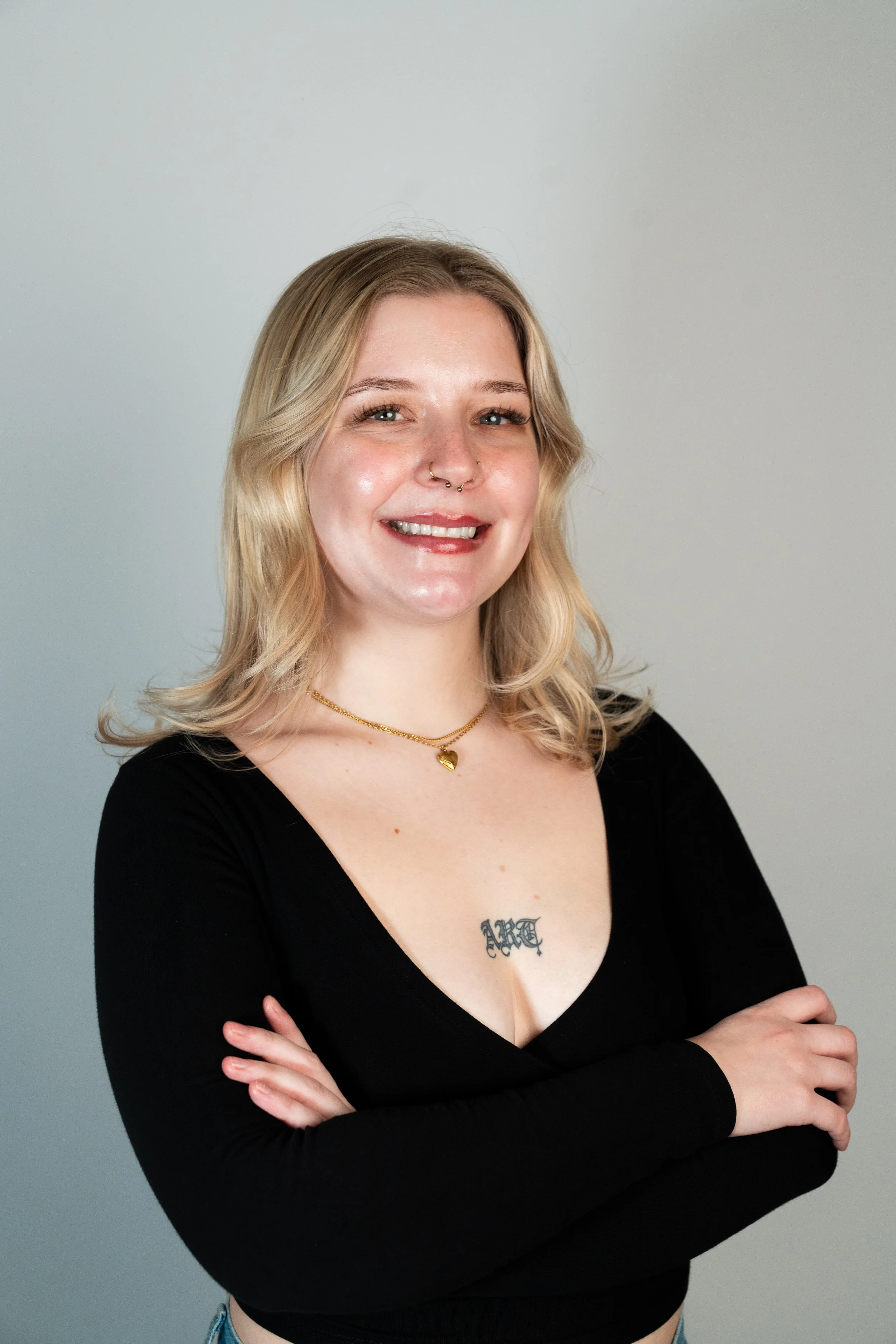 A woman with shoulder-length wavy blonde hair, wearing a black sleeveless top, smiling with red lipstick, showing a tattoo of a sun with a face on her upper arm and a line drawing of clouds surrounding the sun, against a plain white background.