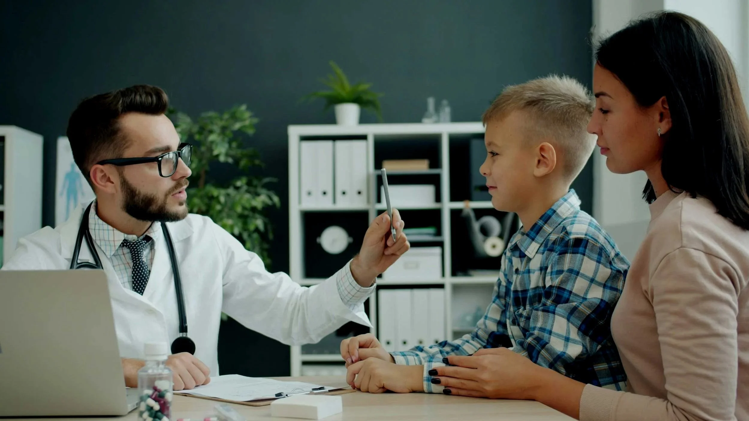 A doctor in glasses and a white coat is speaking to a woman and a young boy in a medical office. The doctor is holding a pen and gesturing towards the boy. The woman and boy are seated at a desk with the woman holding the boy's hand. The background shows shelving with plants and files.