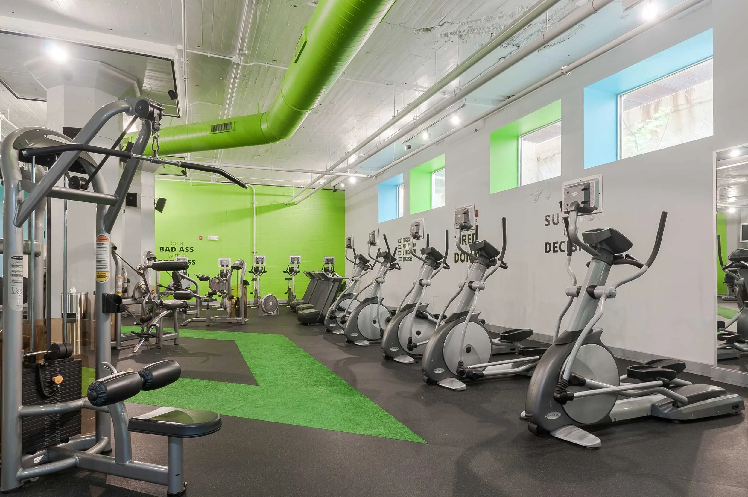 Interior view of a modern gym with cardio equipment, including ellipticals and stationary bikes, featuring green and white walls with motivational messages, and windows letting in natural light.