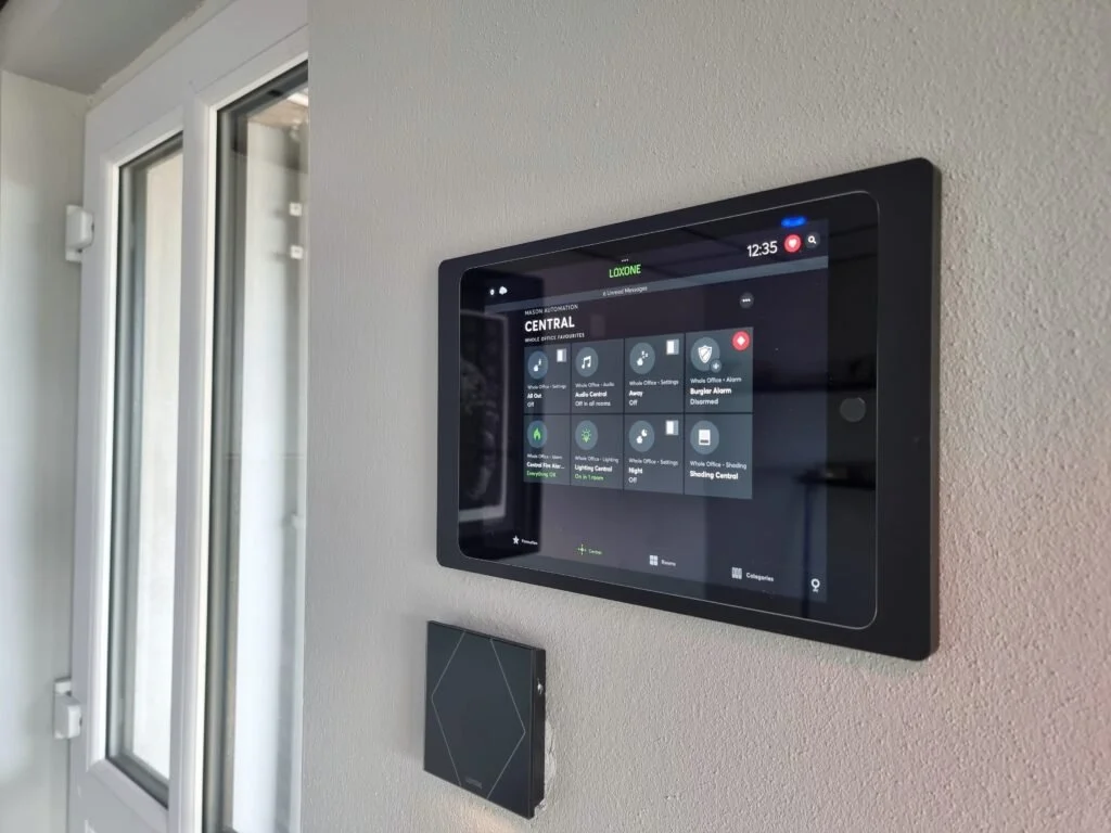 A touchscreen control panel mounted on a wall inside a house, displaying smart home automation options.