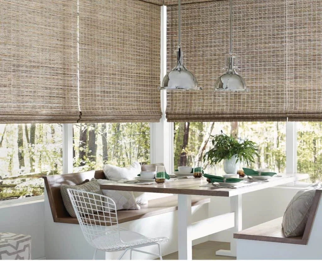 A cozy dining nook with a wooden table set for a meal, surrounded by white and wooden chairs, with large windows showing a forest outside, brown woven blinds, and pendant lights hanging overhead.