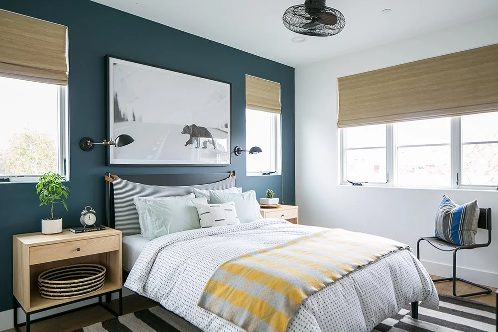 A modern bedroom with a blue accent wall behind the bed, featuring a large framed bear painting, two windows with beige blinds, a wooden nightstand with a plant and alarm clock, a bed with white and striped bedding, a chair with a pillow, and a ceiling fan.