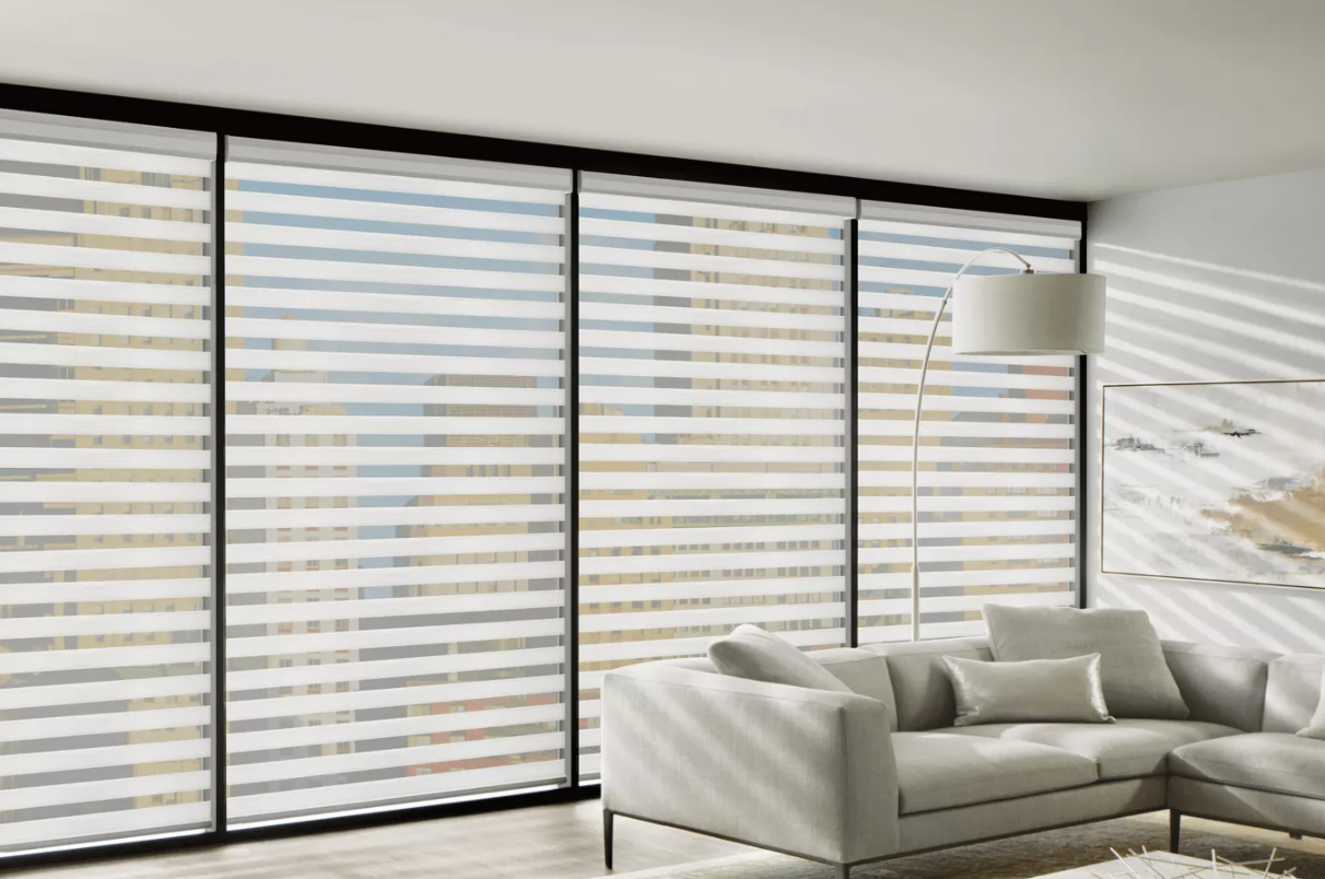 Modern living room with large windows featuring horizontal blinds, a white sectional sofa with pillows, a tall arched floor lamp, and a framed abstract painting on the wall, with city buildings visible through the windows.