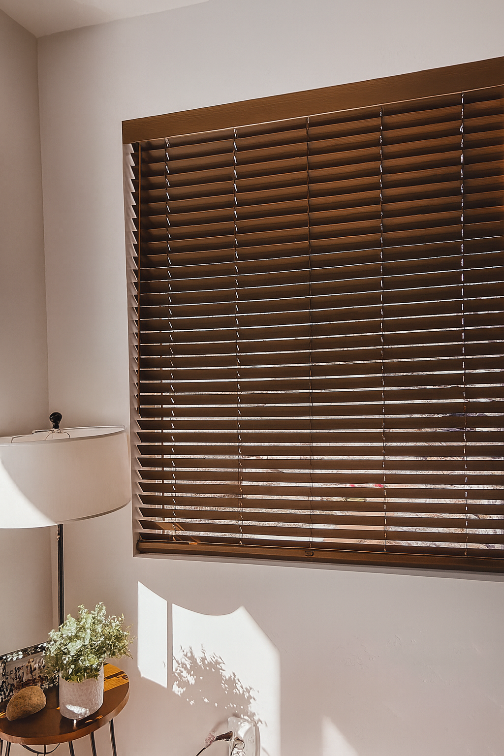 A room with a large window covered by horizontal wooden blinds, a white wall, a corner of a white lamp, and a small round wooden table with a potted plant, a rock, and a white object.