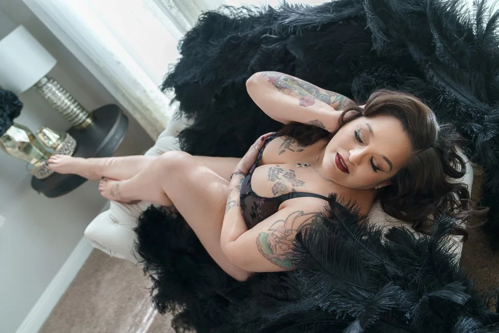 Tattooed woman in black lace lingerie reclines on a shaggy black rug, eyes closed and arms behind her head, creating a sultry, relaxed mood.