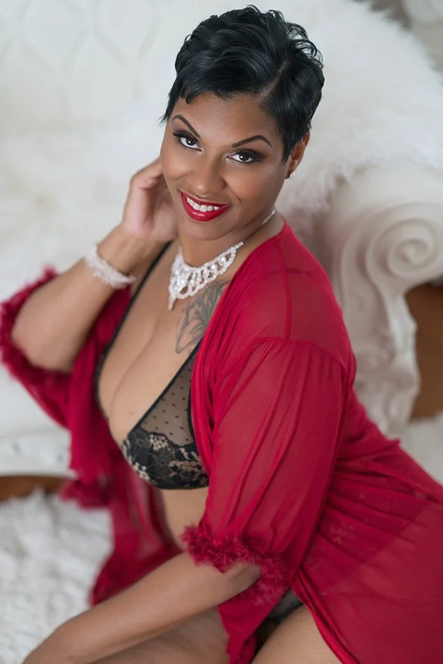 Smiling woman with short dark hair poses in a red robe and black lingerie, wearing jewelry, seated on a plush bed in a glamorous portrait.