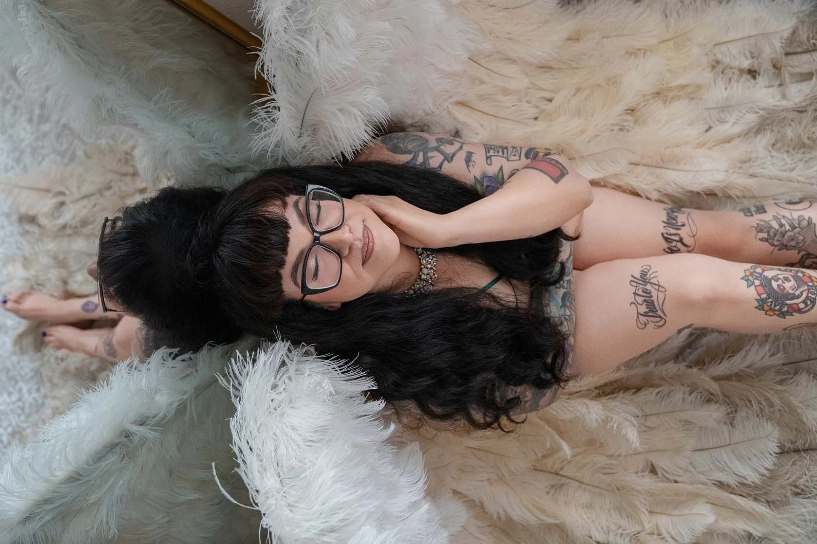 Smiling tattooed woman with long dark hair and glasses reclines on a plush white fur chair, creating a cozy, playful portrait.